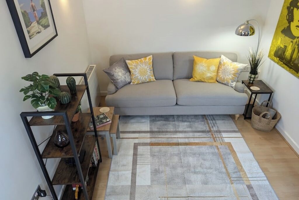 Ein Wohnzimmer mit grauem Sofa, gelb-weißen Kissen, einem gemusterten Teppich, einem kleinen Beistelltisch, einer Stehlampe und einem Bücherregal mit Pflanzen.