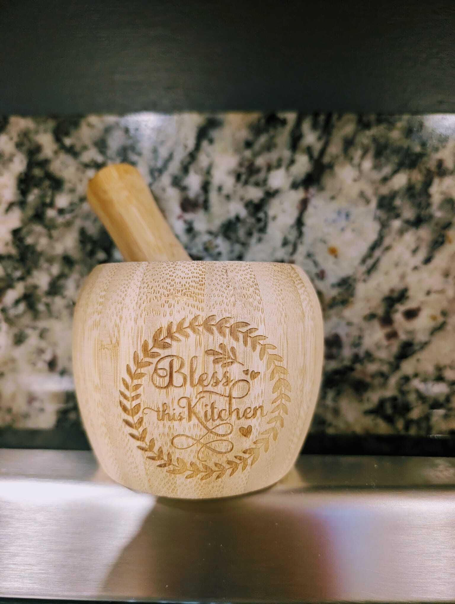 A wooden kitchen utensil with a handle and a bowl, featuring an engraved text "Bless this Kitchen" surrounded by a laurel wreath design.
