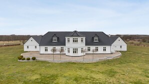 Une grande maison blanche à deux étages, dotée d’une architecture symétrique, avec une partie centrale agrémentée d’un balcon et deux ailes de chaque côté.