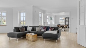 Un salon spacieux avec un canapé d’angle gris, une commode en bois et, au fond, un coin repas.