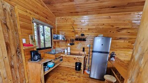 Une petite cuisine en bois, intégrée dans une cabane, avec un réfrigérateur encastré, des étagères en bois et une fenêtre donnant sur la verdure.