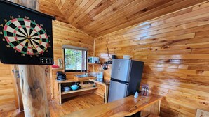 L’intérieur d’un chalet en bois comprend un coin cuisine, un jeu de fléchettes et une fenêtre donnant sur un paysage verdoyant.