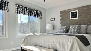 Une chambre à coucher avec un lit, une table de chevet, une lampe et une vue sur les arbres par la fenêtre.