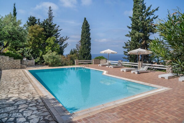 Una piscina rectangular con borde de piedra, rodeada de sillones de descanso y sombrillas, con árboles y vistas al mar al fondo.