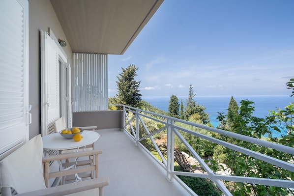 A balcony with a table and chairs, a railing, and a view of the sea and greenery.