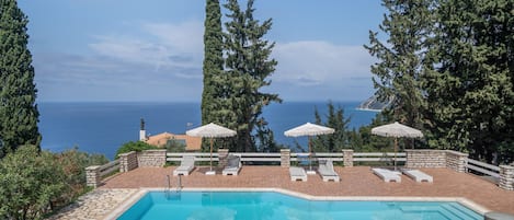 A pool with lounge chairs and umbrellas, surrounded by stone walls and trees, overlooking a body of water.