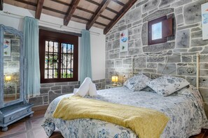 A bedroom with a stone wall, a bed with floral bedding, a window with curtains, and a small table with a lamp. - Holiday Home 'Laura' with Shared Pool, Wi-Fi and Air Conditioning (Conil de la Frontera)