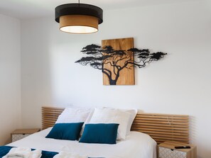 A bedroom with a bed, pillows, a wooden headboard, a wall-mounted artwork of a tree, and a hanging light fixture.