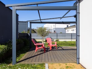 A covered outdoor seating area with two red chairs, a wooden deck, and a view of buildings in the background.