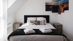 A modern bedroom with a sloped ceiling, a large bed with multiple pillows, bedside tables, and a set of three landscape paintings on the wall.