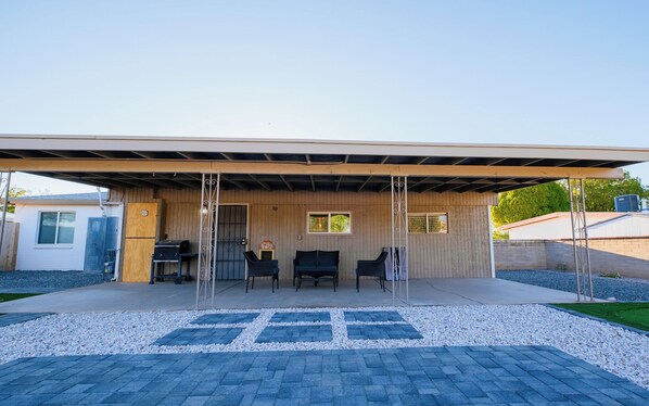 A covered outdoor seating area with a wooden deck, two windows, and a grill.