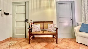 Ein Zimmer mit einer hölzernen Bank mit blumigen Kissen, zwei Türen, einem weißen Sofa mit einem blauen Kissen und einem gemusterten Teppich auf dem Boden.