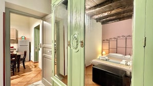 Ein Zimmer mit einem Bett, einem Nachttisch und einem Spiegel. Eine Tür führt zu einer Küche mit einem Kühlschrank und einem Esstisch.