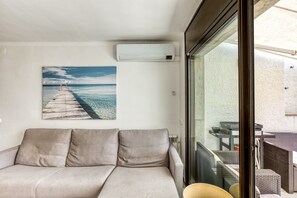 Una habitación con sofá color beige, un cuadro de un muelle, un equipo de aire acondicionado y una puerta de cristal que da a un espacio exterior con mobiliario de mimbre.