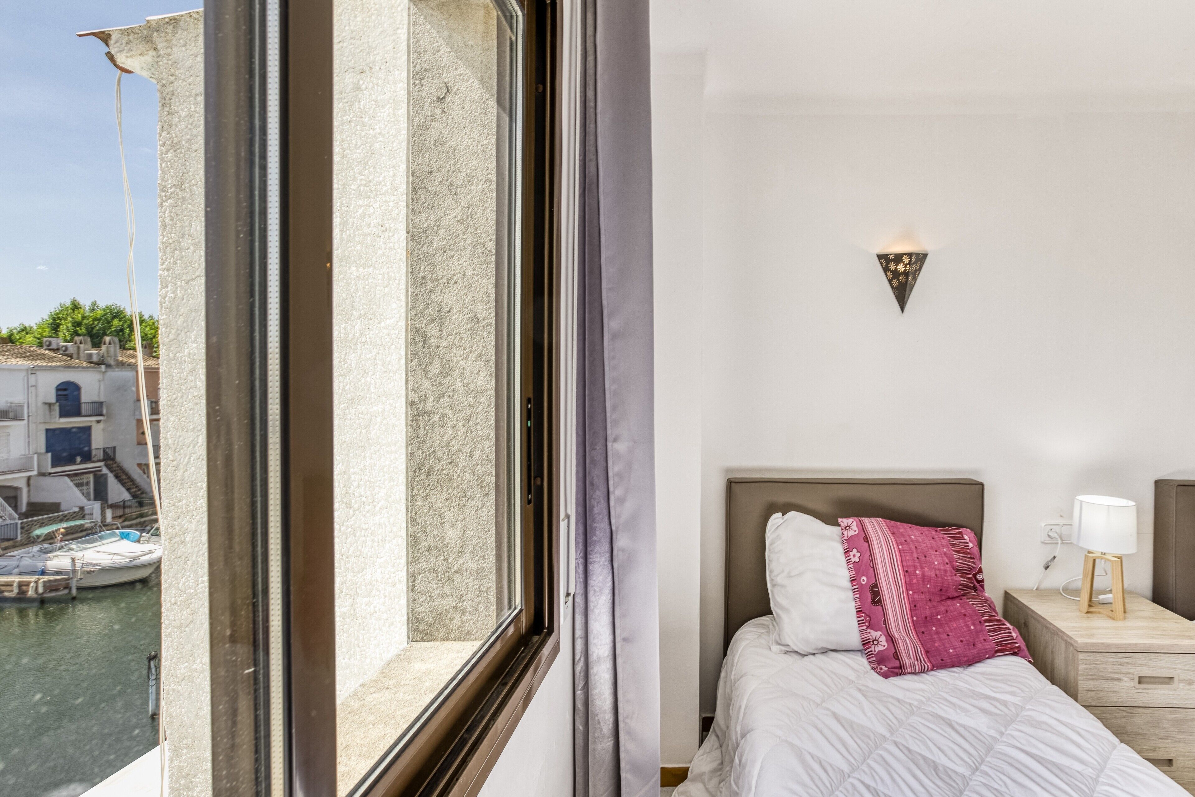 Une chambre d’hôtel avec une grande fenêtre, un lit avec une literie blanche, un oreiller à motifs roses, une table de chevet avec une lampe et un luminaire fixé au mur.