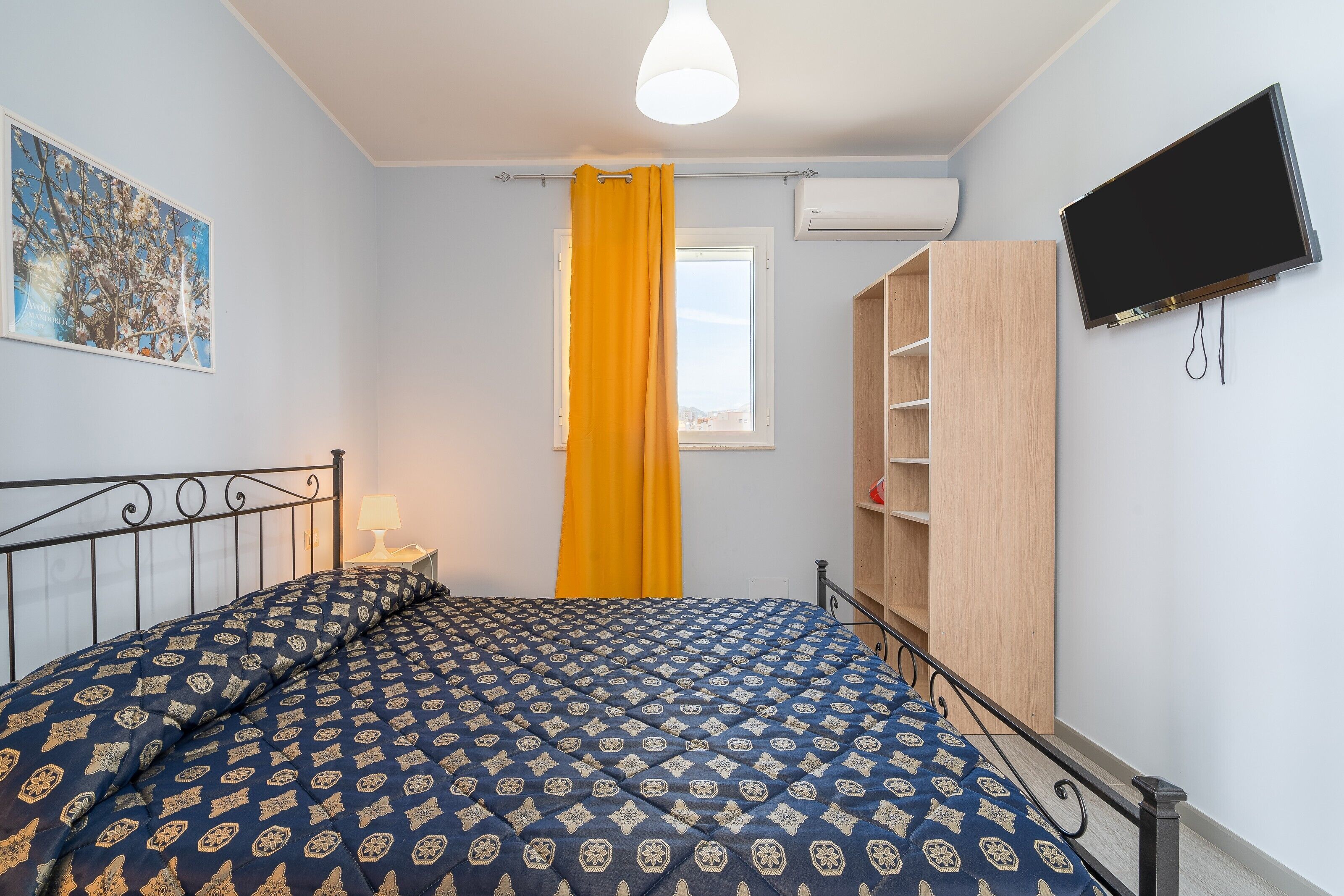 A bedroom with a bed, a bedside table, a TV, a window with curtains, and a wall-mounted shelf.