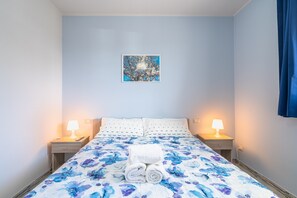 A bedroom with a bed, two bedside lamps, a wall-mounted picture, and a neatly folded towel on the bed.