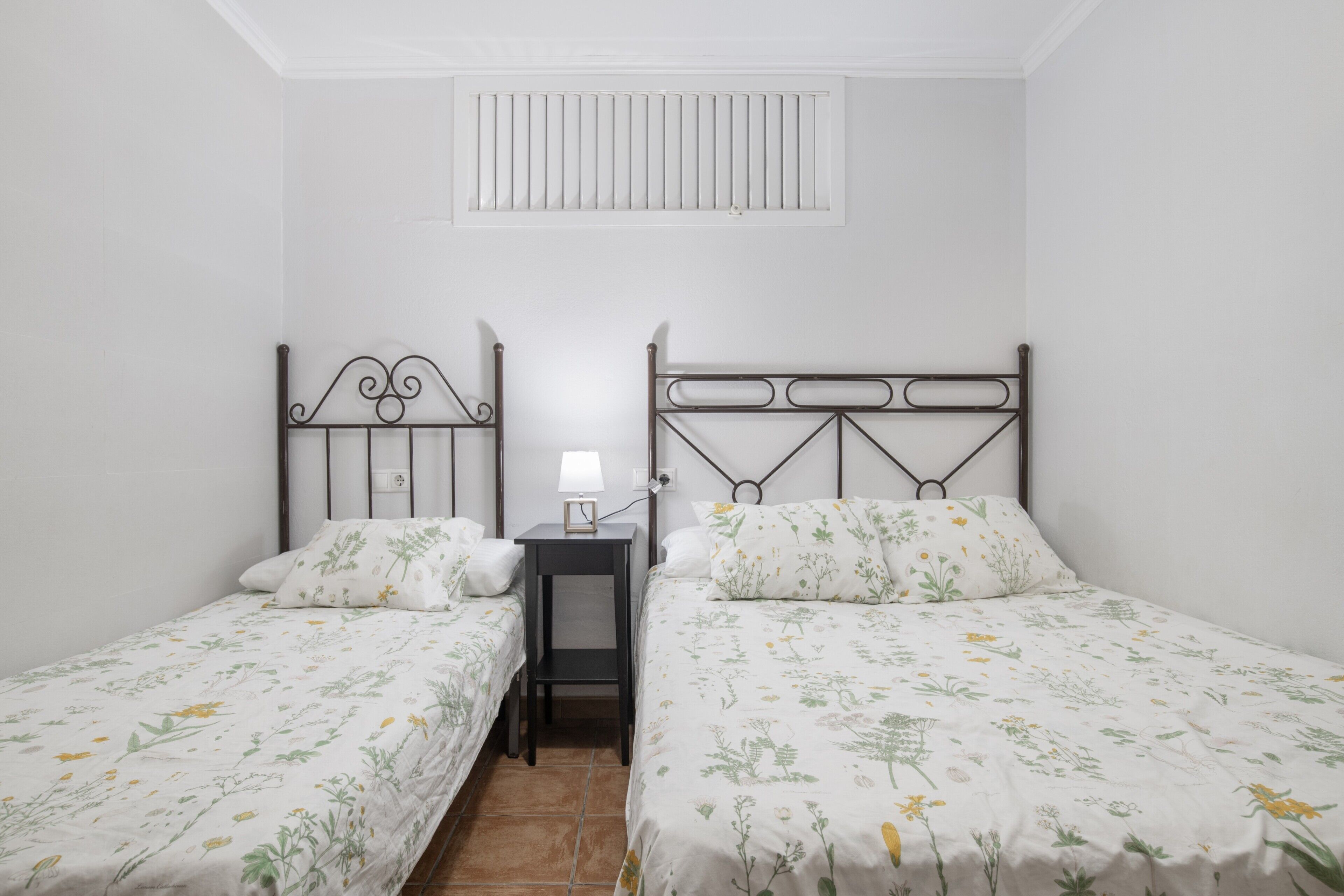 A bedroom with two single beds, a nightstand, a lamp, and a window with blinds.