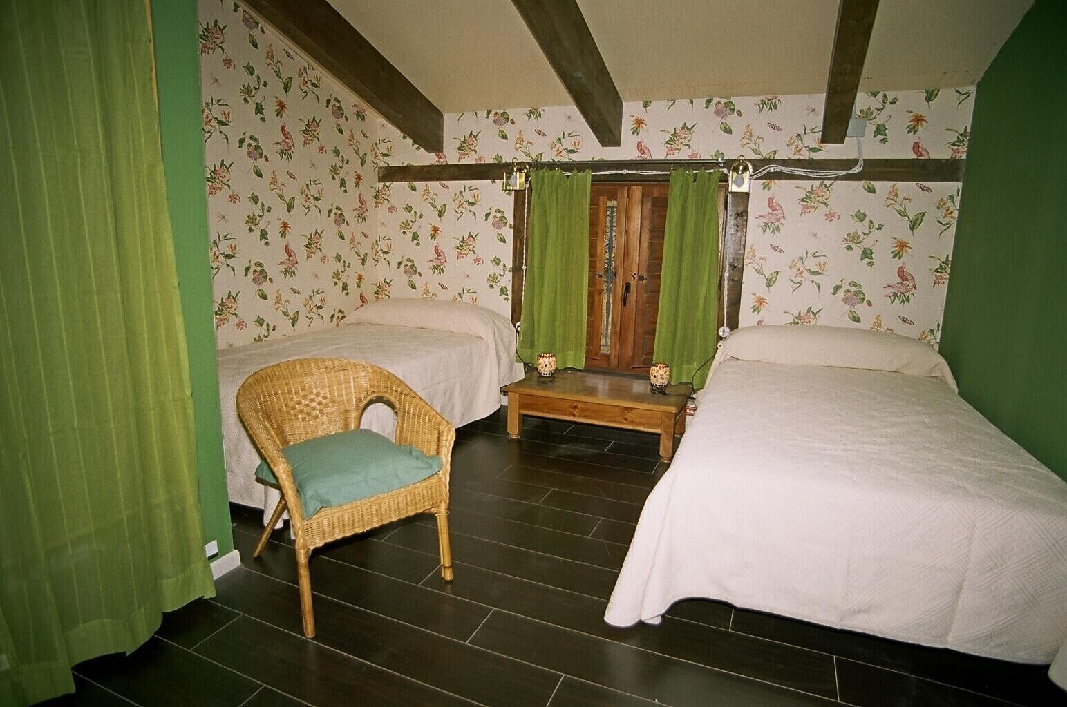 Una habitación con dos camas, una silla de mimbre, una mesa de madera y cortinas verdes.