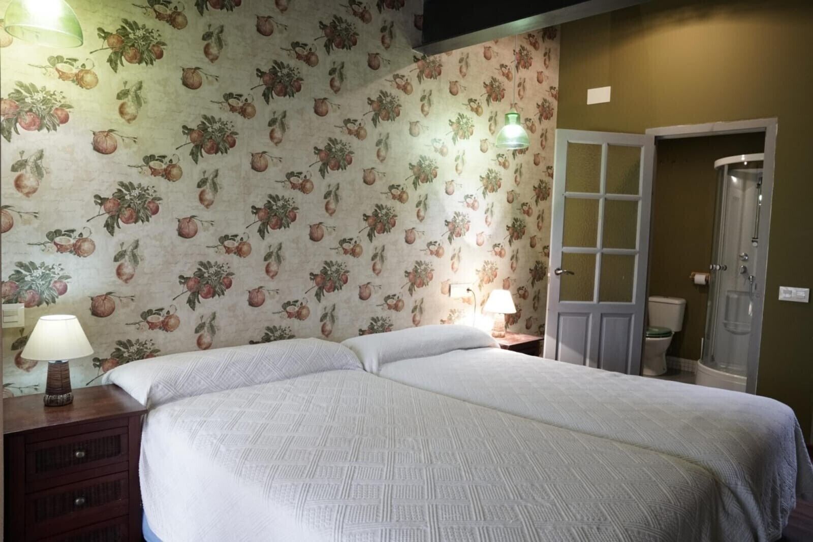 Un dormitorio con dos camas, una mesita de noche con lámpara, una puerta a un ba ño y una ducha.