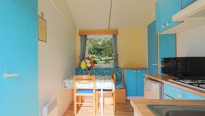A compact kitchen with a dining area, a microwave, a small refrigerator, and a window with curtains. - Mobile Home 'Bikini' with Private Terrace and Private Garden (Plouha)