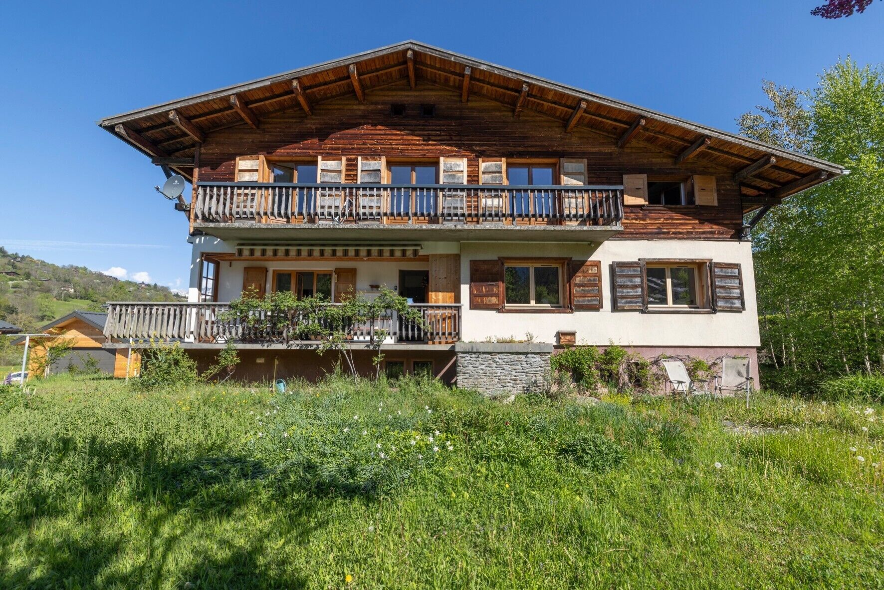 A two-story wooden chalet with a balcony, stone foundation, and a view of the surrounding landscape.