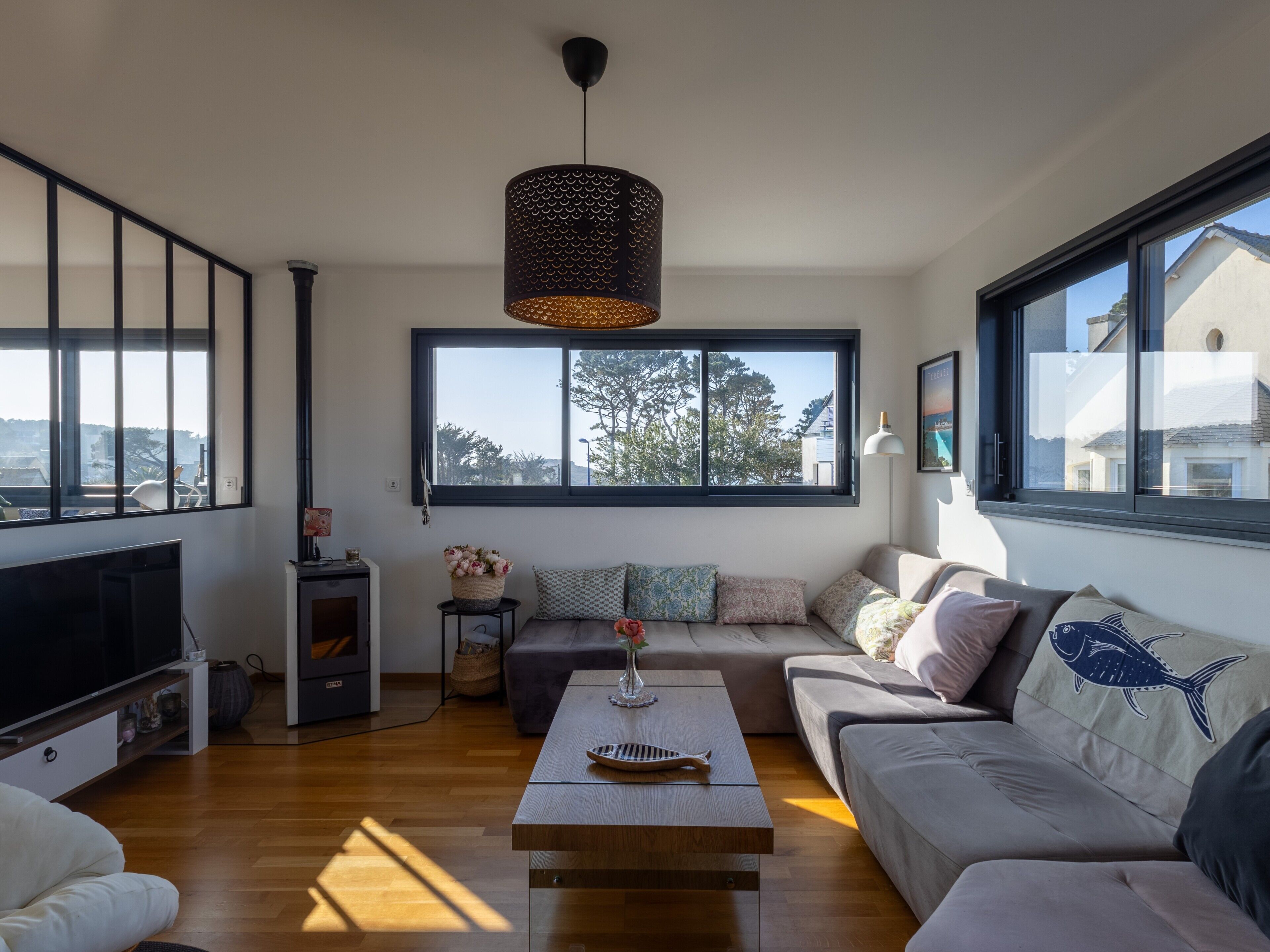 A modern living room with a large window, a flat-screen TV, a wooden coffee table, and a sofa with patterned cushions.