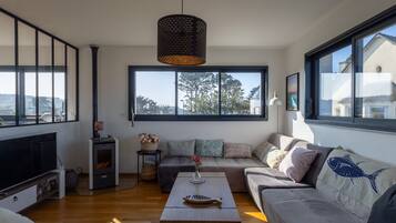 A modern living room with a large window, a flat-screen TV, a wooden coffee table, and a sofa with patterned cushions.