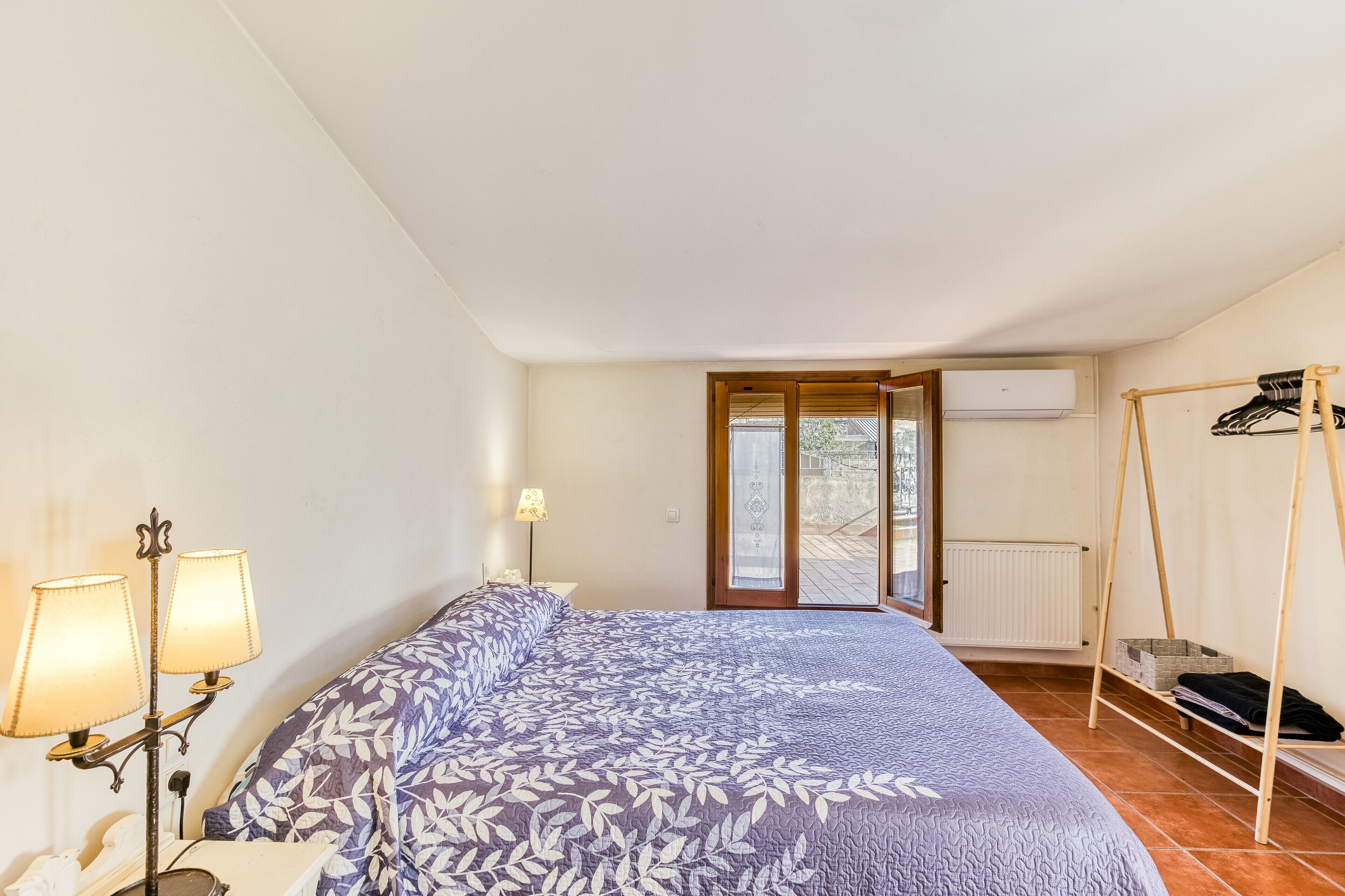 Ein Schlafzimmer mit einem Bett, zwei Nachttischlampen, einem Schaukelstuhl aus Holz und Blick durch eine Tür auf einen Außenbereich.