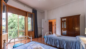 A bedroom with two beds, a wooden wardrobe, a window with a view of trees, and a door leading to another room.