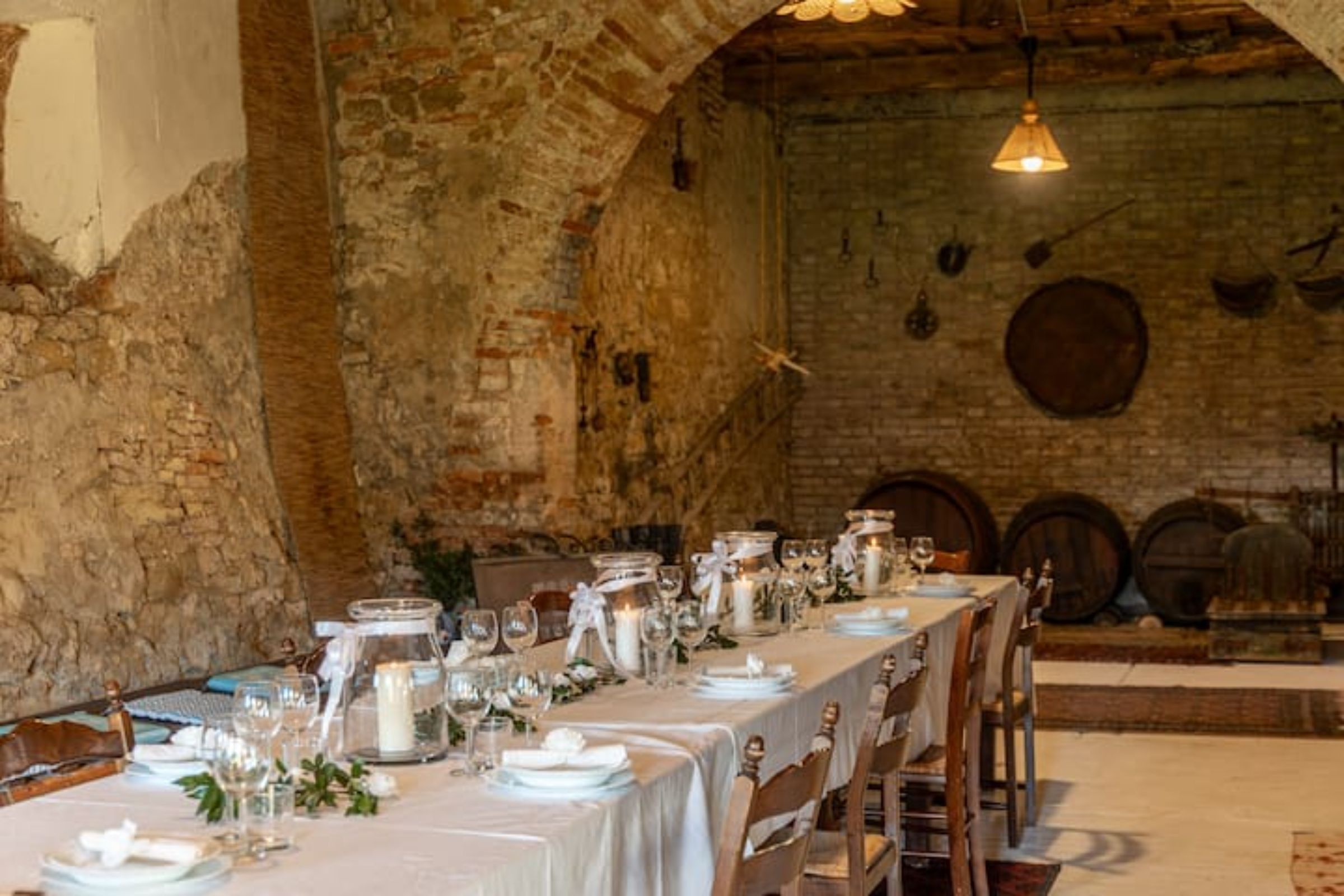 Un lungo tavolo imbandito con tovaglie bianche, bicchieri e candele, in una rustica stanza con pareti in mattoni e botti di legno sullo sfondo.