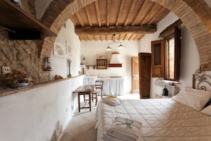 Een slaapkamer met een stenen muur, houten balken in het plafond, een bed met een sprei, een nachtkastje en een raam.