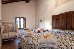 A bedroom with a crib, a bed with patterned bedding, a wooden wardrobe, and a window.
