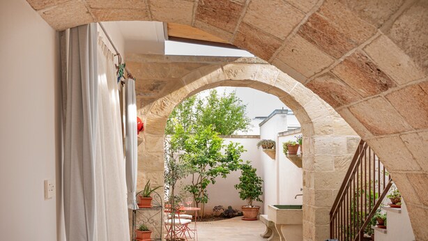 Un corredor de piedra en arco con plantas en macetas y una mesa con sillas.