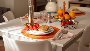 Una mesa de comedor con mantel blanco, una botella, un tazón de tomates, un plato con un panecillo y una cesta de frutas.