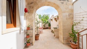 Un arco de piedra que da acceso a un patio con plantas en macetas, una mesa pequeña y una pared blanca.