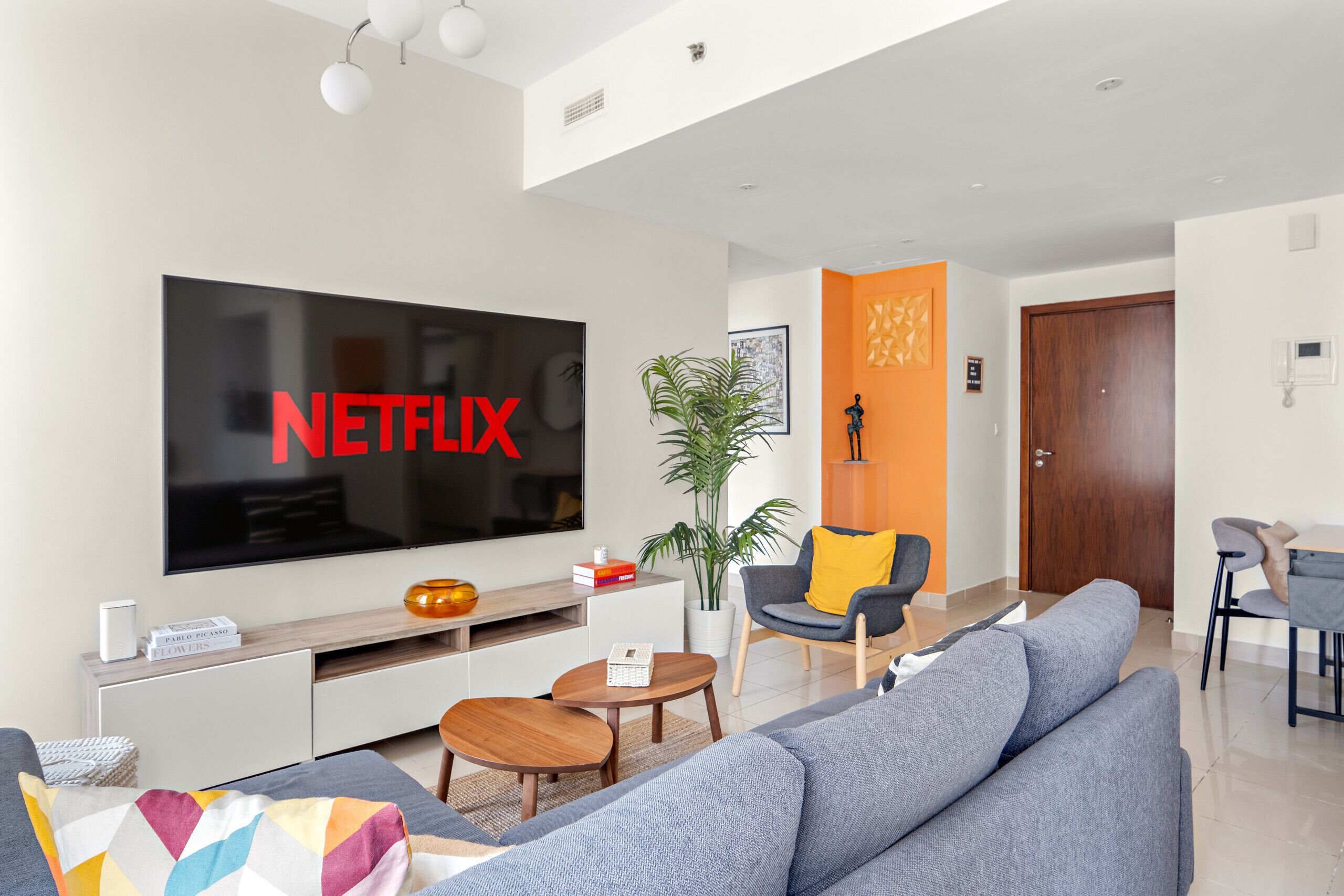 A modern living room with a large flat-screen TV, a sofa, a coffee table, and a dining area with a table and chairs.