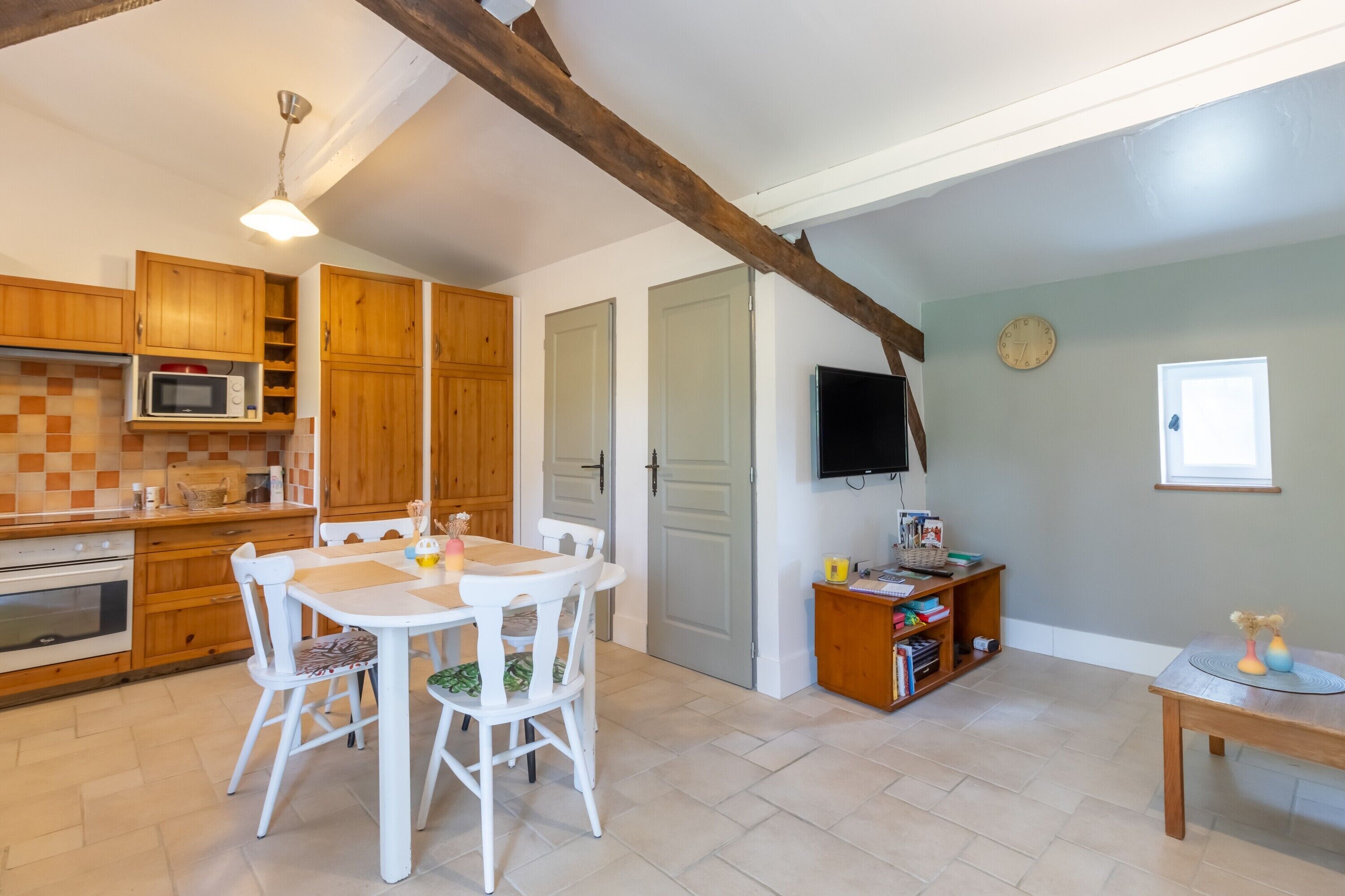 A kitchen with wooden cabinets, a microwave, a dining table with chairs, a television, and a bookshelf.