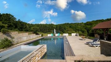 Une piscine rectangulaire, avec un mur en pierre d’un côté et une terrasse en bois de l’autre. On y trouve des chaises longues et une petite table.