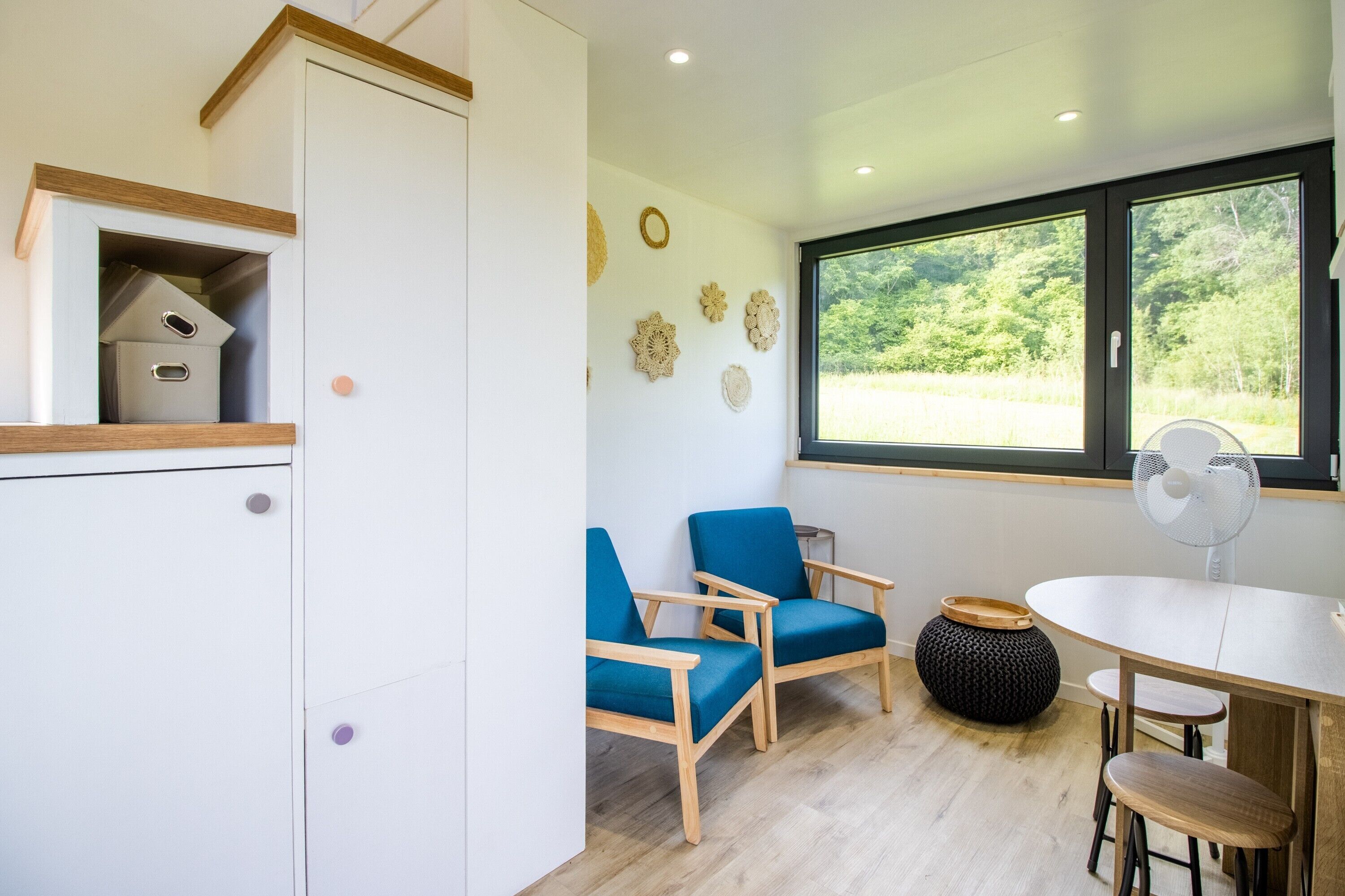 A room with a wooden cabinet, two blue chairs, a round table, a fan, and a window with a view of greenery.