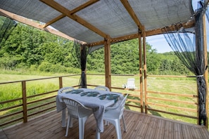 A covered outdoor area with a table and chairs, surrounded by a wooden deck and a view of a grassy field.