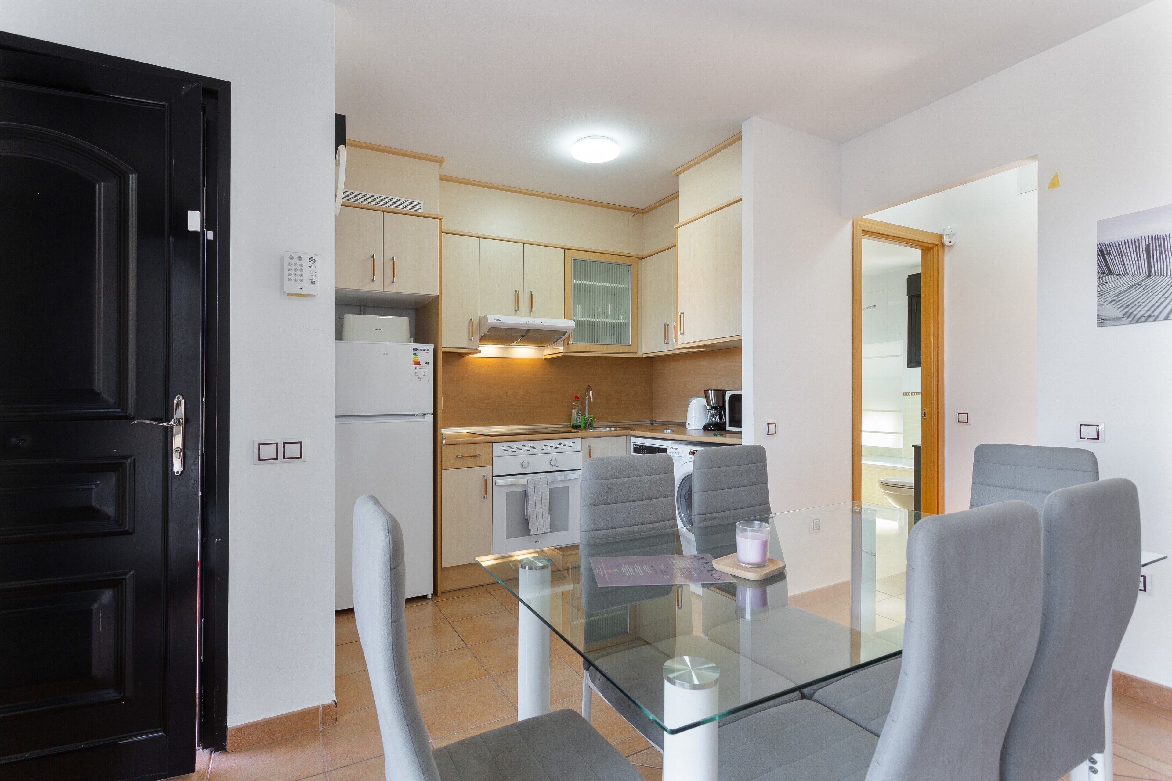A modern kitchen with a glass dining table, grey chairs, and a black door.