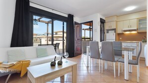 A modern living room with a sofa, a glass dining table, and a kitchen area with cabinets and a stove. - Vacation 'Punta Calma' with Sea View, Shared Pool and Wi-Fi (Majanicho)