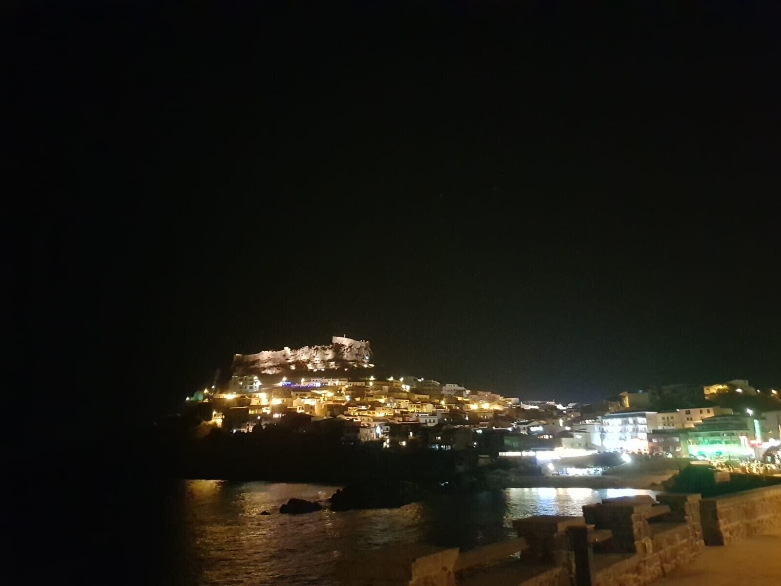 Una cittadina costiera di notte con edifici illuminati, un'imponente struttura collinare e un lungomare.