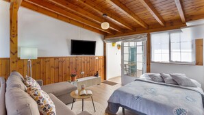 Una stanza con soffitto in legno, un letto con copripiumino decorato, un divano con cuscini ornamentali, un tavolino con un libro e una lampada, e un televisore fissato alla parete.