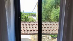 A room with a bed, a window with curtains, and a view of a tiled roof and greenery outside. - Apartment 'Casa Rural Maximón Azul' with Sea Views, Wi-Fi and Air Conditioning (Los Caños de Meca)