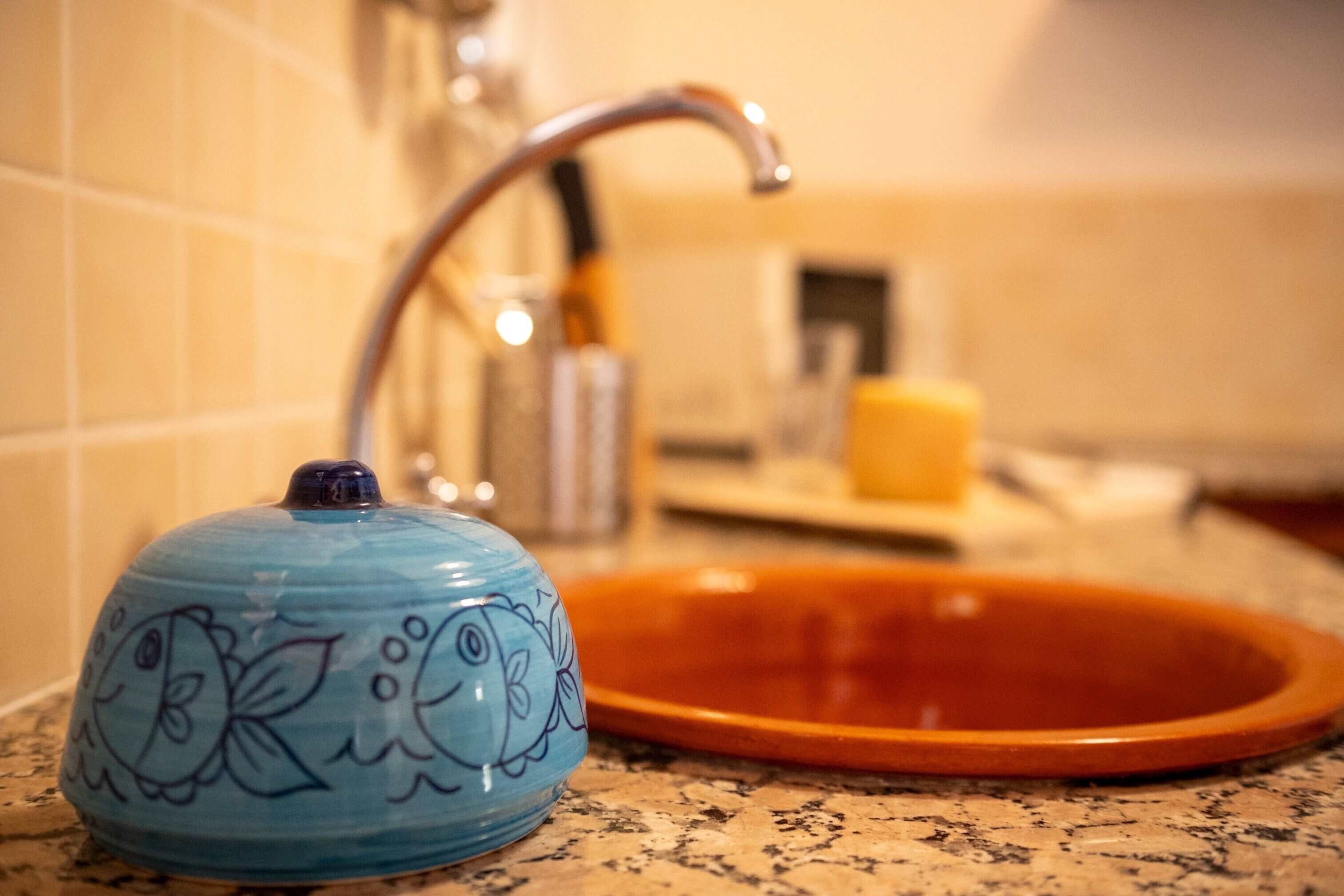 A blue ceramic kettle with floral designs on a kitchen counter, a stainless steel faucet, and an orange sink.