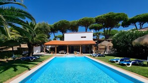 A rectangular swimming pool with clear blue water, surrounded by a white building with a red roof, and trees in the background. - Apartment 'Casa Rural Maximón Azul' with Sea Views, Wi-Fi and Air Conditioning (Los Caños de Meca)