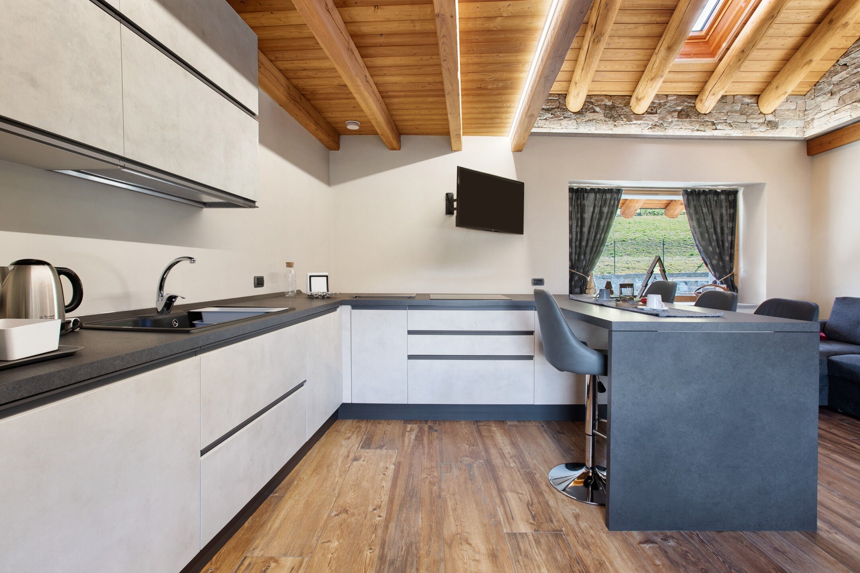 Una cucina moderna con isola integrata, travi in legno al soffitto e vista su un paesaggio verde attraverso una finestra.