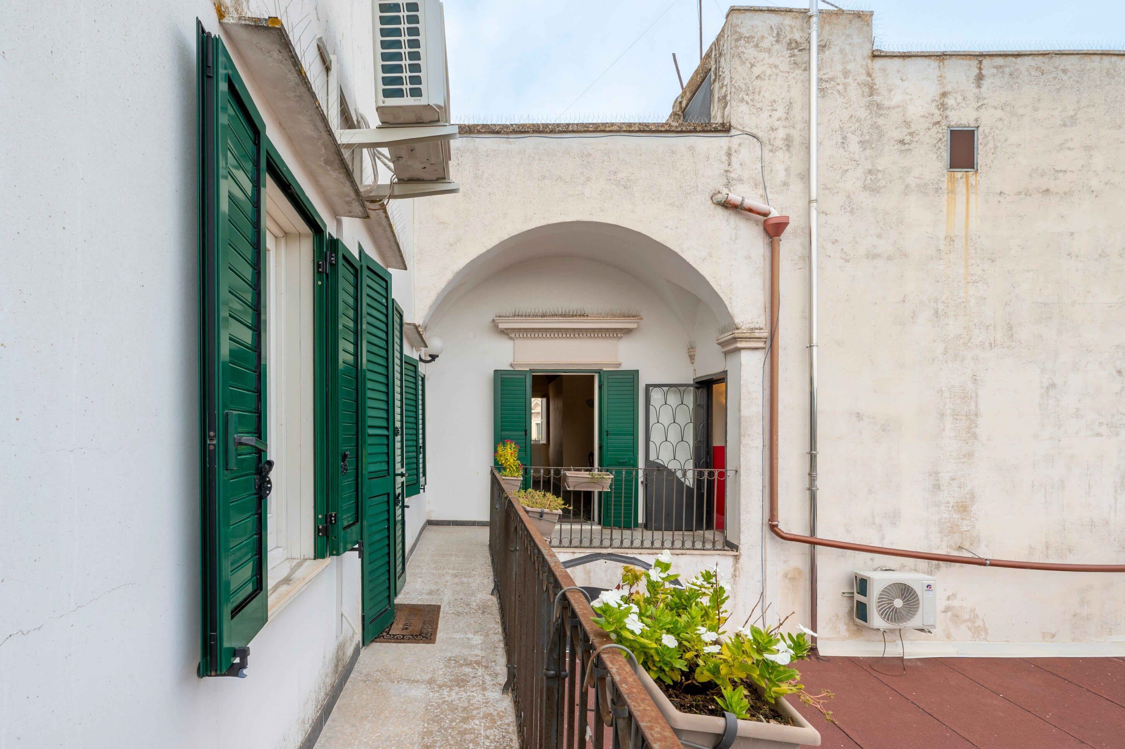 Un vialetto stretto con un edificio bianco, persiane verdi e un piccolo balcone con piante in vaso.